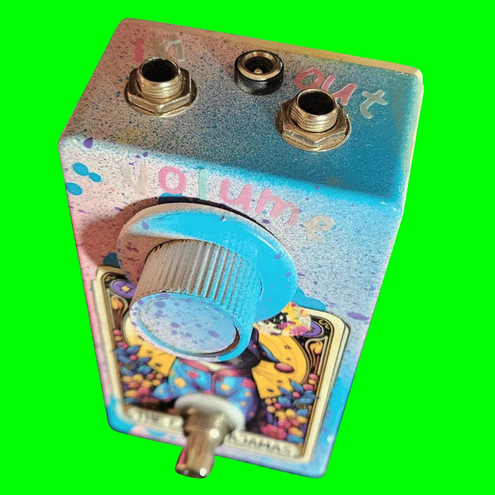 Top-down view of the enclosure: a 1/4-inch jack sits at each corner of the top edge, hand-lettered "in" on the left and "out" on the right, with a barrel DC power jack centered between them. Pastel letters spelling "volume" arc above the large light-blue knurled knob.