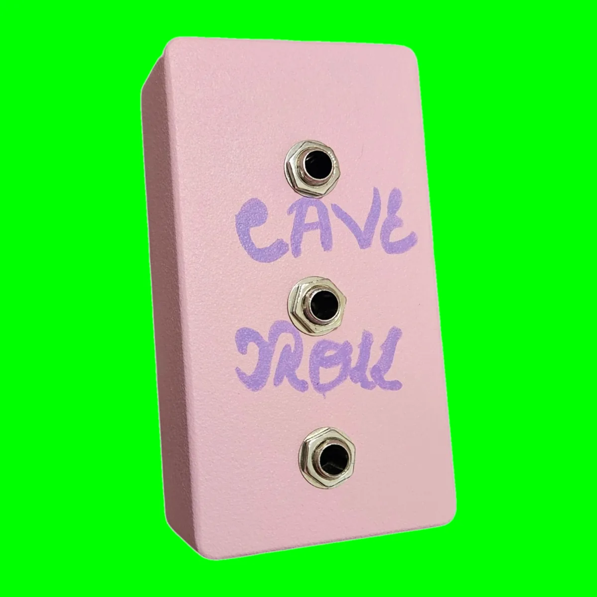 A pale pink pedal photographed at a three-quarter angle. CAVE TROLL is hand-lettered across the face in purple marker in two lines, CAVE above and TROLL below, with three quarter-inch jack sockets running down the face between and below the words, each with a silver hex nut. There are no knobs, no switch and no indicator lamp anywhere on it.