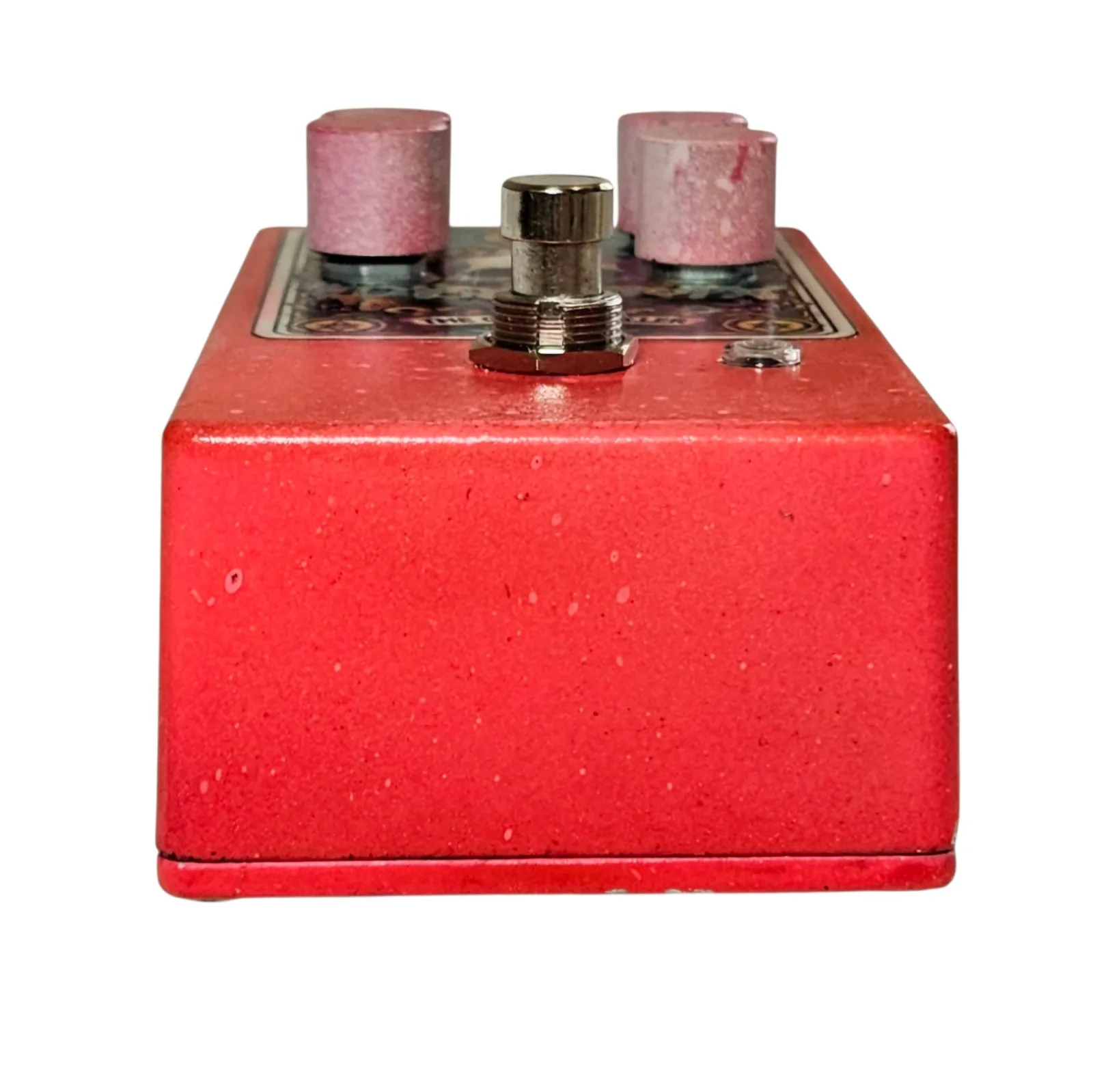 The pedal seen end on from the footswitch side. The red speckled wall of the enclosure fills the frame, with the silver footswitch, two of the pink knobs and the small LED ranged along the face above it.