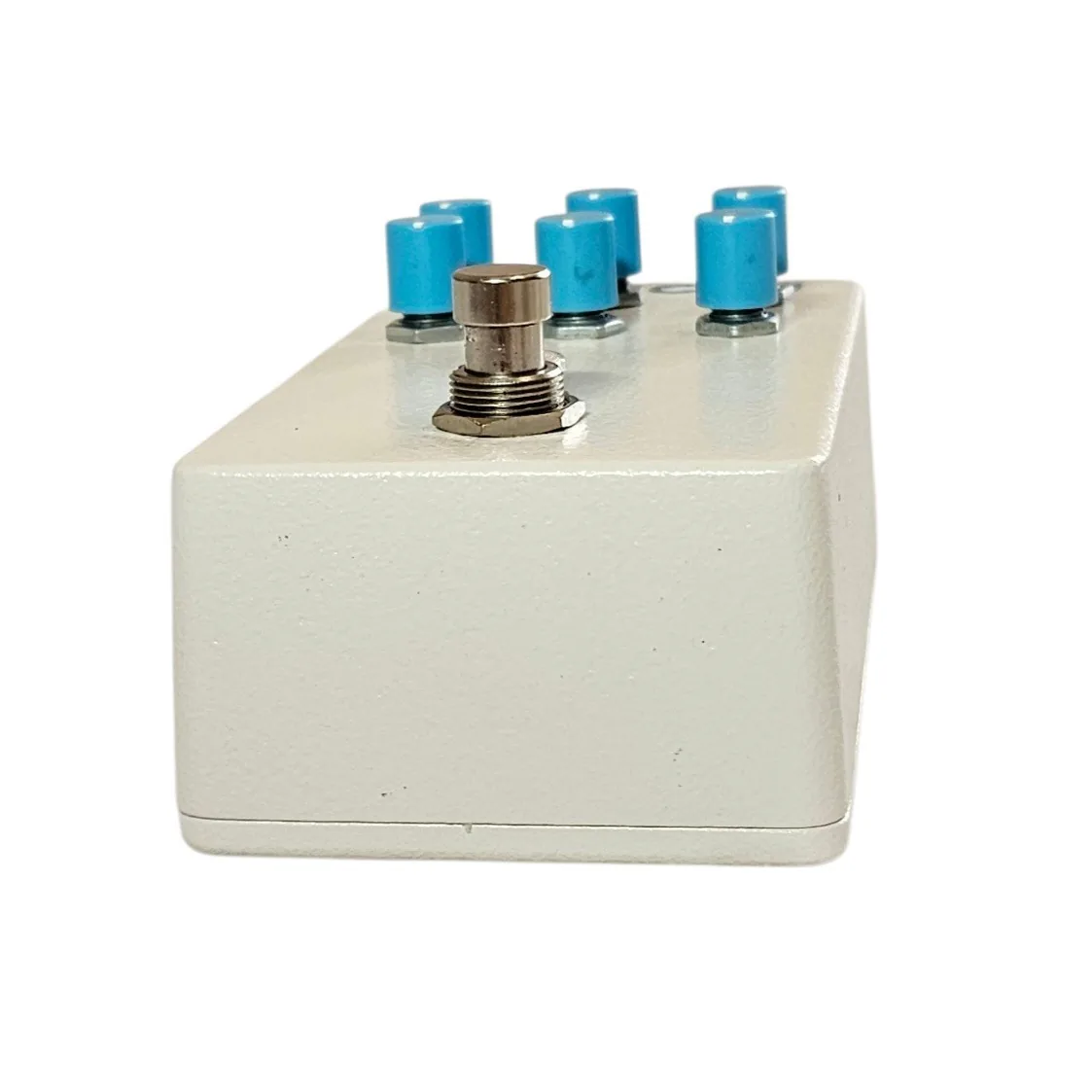Looking down on the pedal from the footswitch end: the six blue knobs stand along the far edge in two rows, the silver footswitch nut is in the foreground, and the plain white wall of the enclosure fills the lower half.