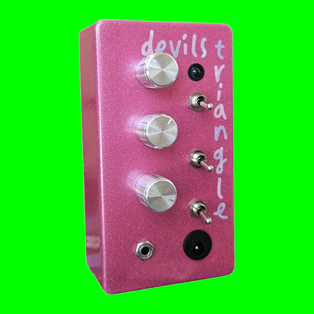 Devil's Triangle - an upright pink sparkle box with three silver knobs down the left, three toggle switches and an LED down the right, devils triangle lettered down the side, a 3.5 mm jack and a DC socket at the bottom