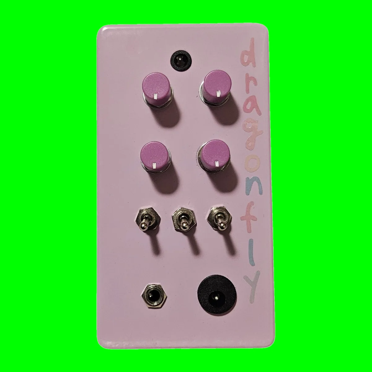 Dragonfly - an upright lilac box with four pink knobs in two rows, three toggle switches below them and a small 3.5 mm jack and DC socket at the bottom, the name in pastel letters down the side, on a chroma green tile