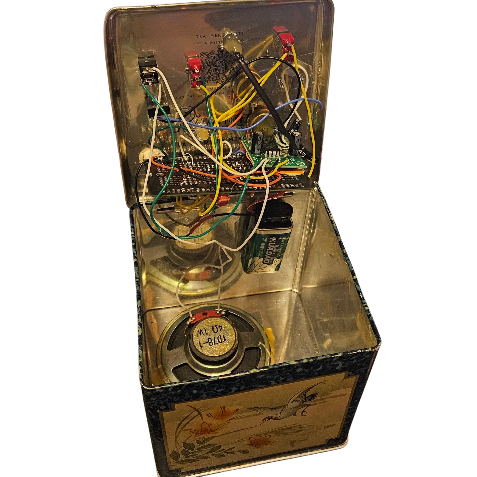 The tin standing open with the lid tipped back. A dark solderable breadboard is mounted inside the lid, crossed by white, yellow, green, orange, blue and black wires, with a small green amplifier module and its electrolytic capacitors soldered on at one end and the red and black bodies of the lid-mounted controls ranged along the top. One thick black lead stands proud of all the others and runs diagonally across the board: that is the touch bend wire. Behind the wiring the tin's polished lining shows its printed tea-merchant royal warrant. In the bottom of the tin a green 9V battery lies beside a round speaker marked TD78-1, 4 ohm 1W, wired back up to the board, and one painted side showing a flying crane is visible at the front.