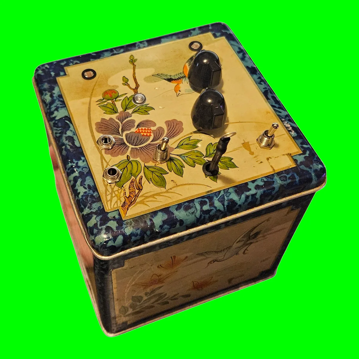 A square tea tin painted with birds and flowers inside a mottled blue border, with two black knobs, two toggle switches, the photocell eyes and a touch probe on a black lead set into the lid