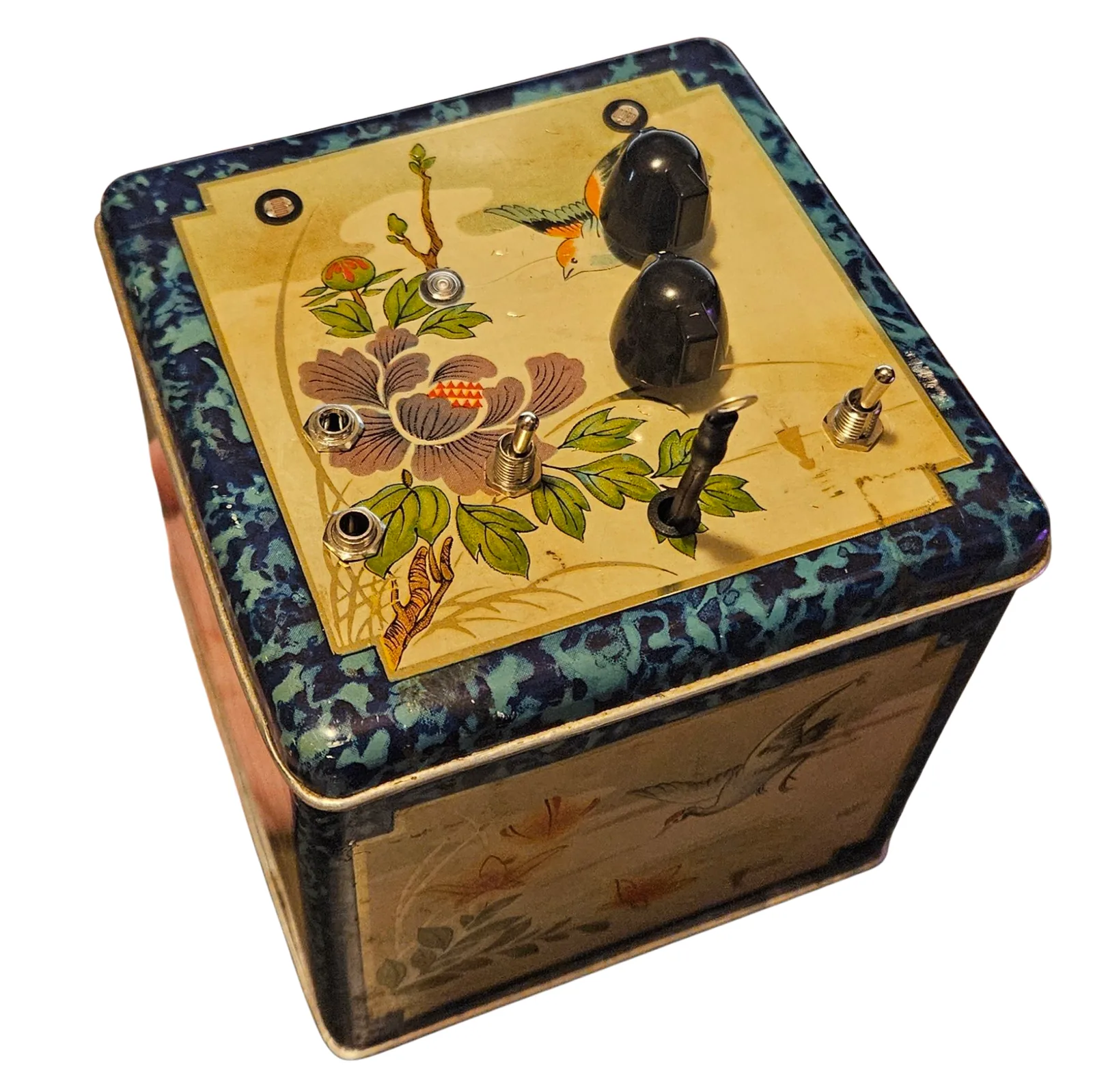 The Duet Deluxe from above and slightly to one side: a square tea tin with a mottled dark blue border and a cream panel painted with a bird, a purple peony and green leaves. Set into the lid are two black knobs, two toggle switches, two small dark photocell windows, a round LED, two chrome jack collars, and a black lead that stands up out of a grommet and ends in a metal ring terminal - the touch probe. One painted side of the tin, showing long-tailed birds, is visible below.