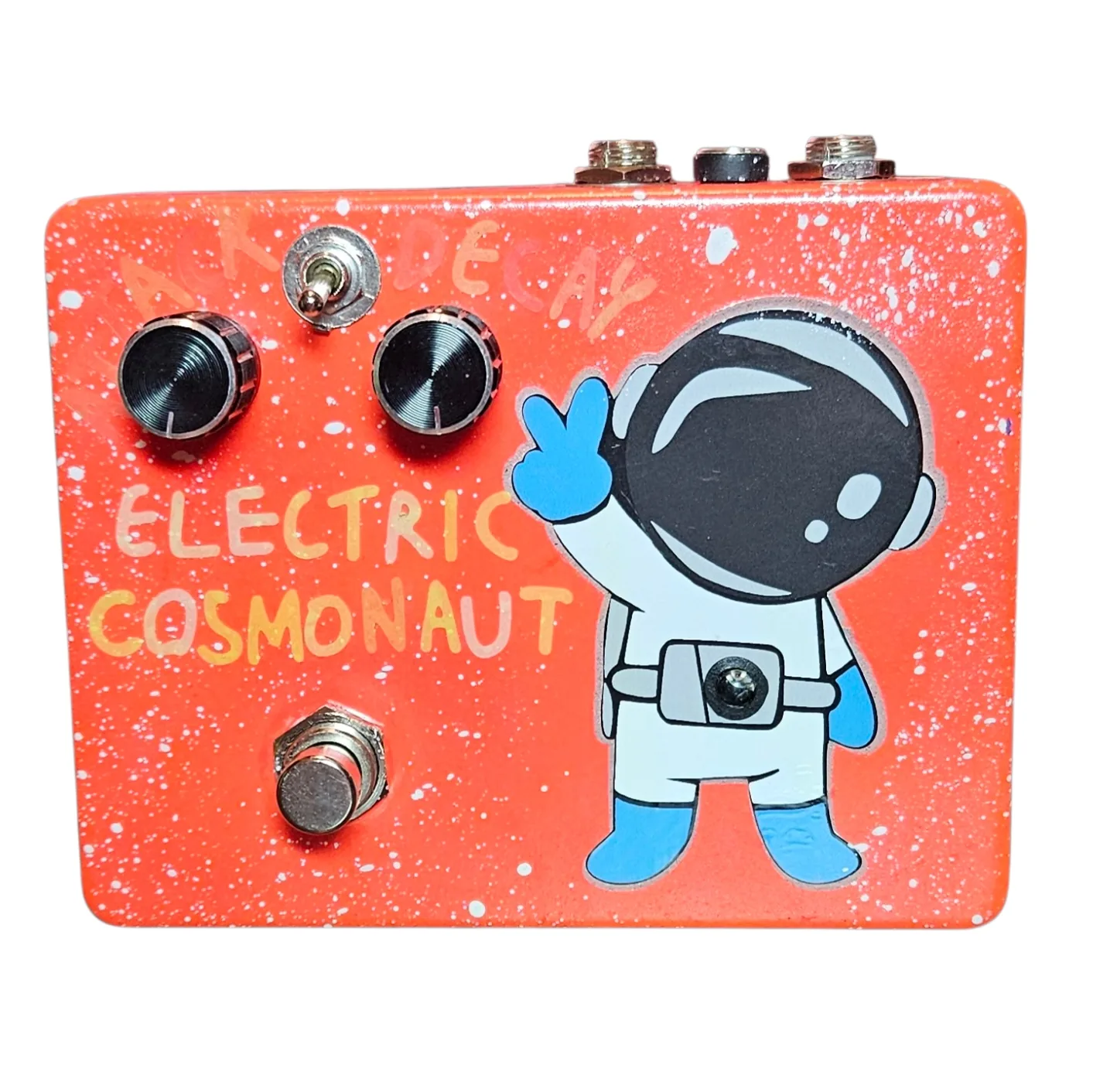 Electric Cosmonaut pedal, front view - orange enclosure flecked with white speckle like a starfield, with engraved Attack and Decay knobs in ribbed cream, a Blend toggle between them, and hand-lettered &ldquo;ELECTRIC COSMONAUT&rdquo; in cream and yellow. A waving cartoon astronaut in a white suit with blue gloves and boots fills the right side, and the pedal's LED is set into the center of his belt buckle.
