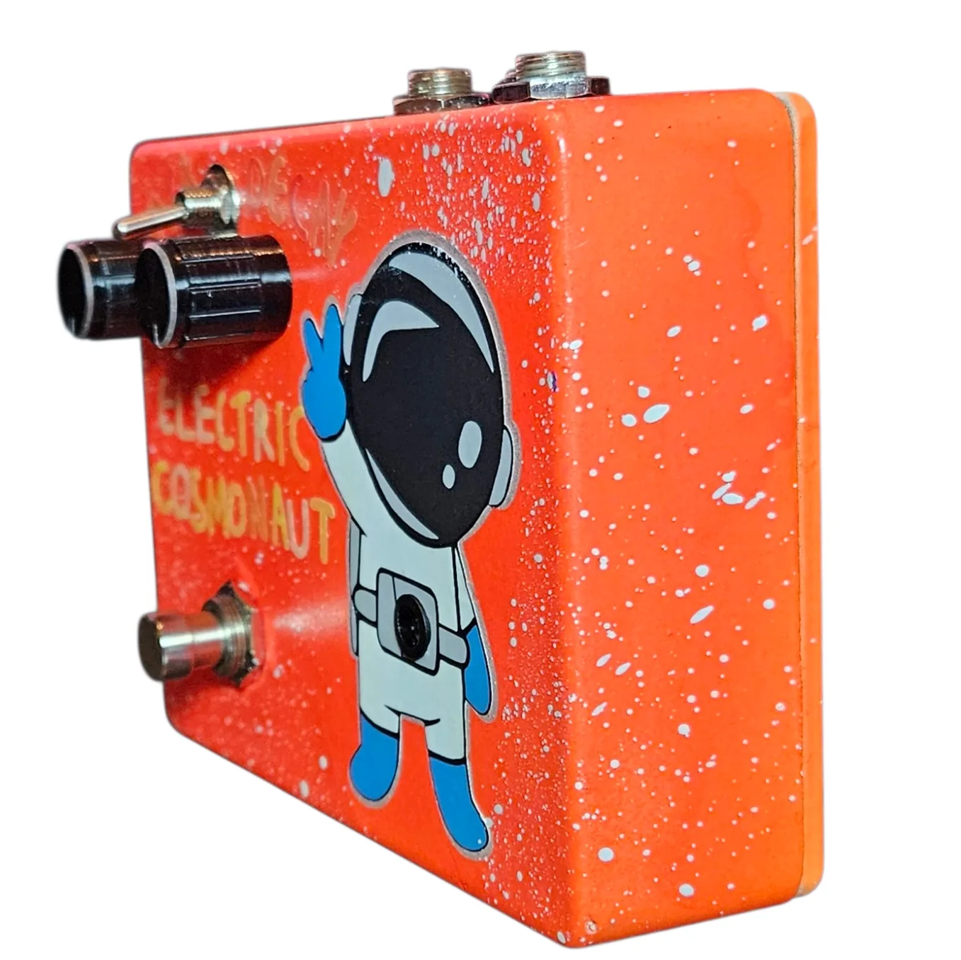 Left side of the pedal: an orange wall flecked with white speckle, showing the two cream knobs and the toggle switch standing out from the face and the footswitch protruding at the bottom. Hand-lettered "ELECTRIC COSMONAUT" runs across the front in cream and yellow beside the waving astronaut.