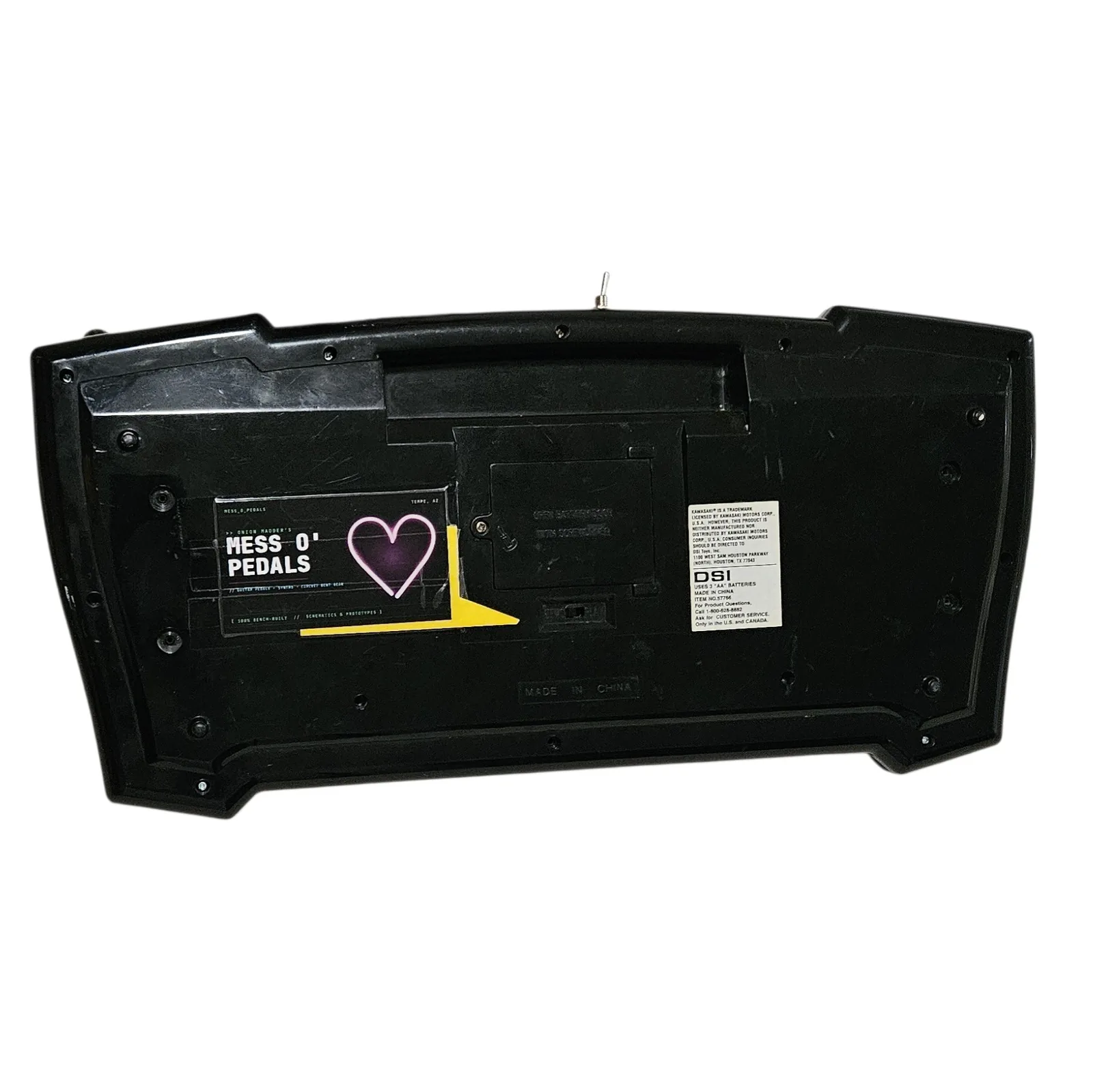 The underside, plain black plastic with the battery door in the middle and MADE IN CHINA molded into it. A black Mess O' Pedals maker sticker is stuck on the left, reading ONION MADDER'S MESS O' PEDALS, Tempe AZ, guitar pedals, synths, circuit bent gear, 100% bench-built, with a purple heart and a yellow arrow. On the right is the original factory label: a Kawasaki trademark notice, DSI Toys of Houston Texas, uses 3 AA batteries, item number 57756.