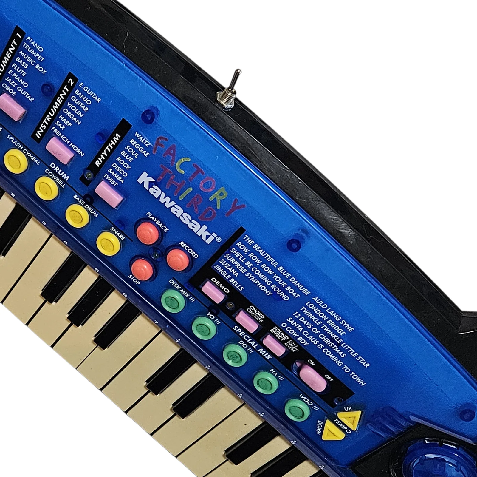 A close angled view across the panel with the hand-lettered FACTORY THIRD filling the middle of the frame, each letter in a different color of marker, sitting directly above the printed Kawasaki logo. One of the added silver toggle switches stands up from the black top edge behind it, and the yellow drum buttons and green SPECIAL MIX buttons run away toward the bottom of the picture.