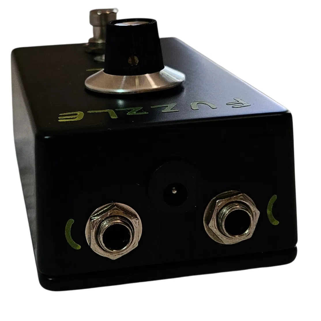 The pedal's top edge straight on, with two silver quarter-inch jack sockets and a recessed barrel power socket set between them. The input is at the left and the output at the right. A curved hand-painted green mark sits beside each jack.