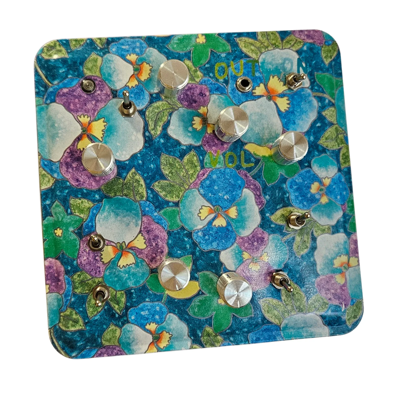 A square floral tin lid, face on and turned slightly, printed edge to edge with pansies in blue, purple, white and green with yellow centers, against white. Set into it are six brass knurled knobs, six brass toggle switches, a small round indicator lamp at the top left, unlit, and a brass jack socket near the top right. Three labels are hand-lettered on the lid in green: OUT beside the jack, ON beside the toggle in the top right corner, and VOL beneath the knob at the center.