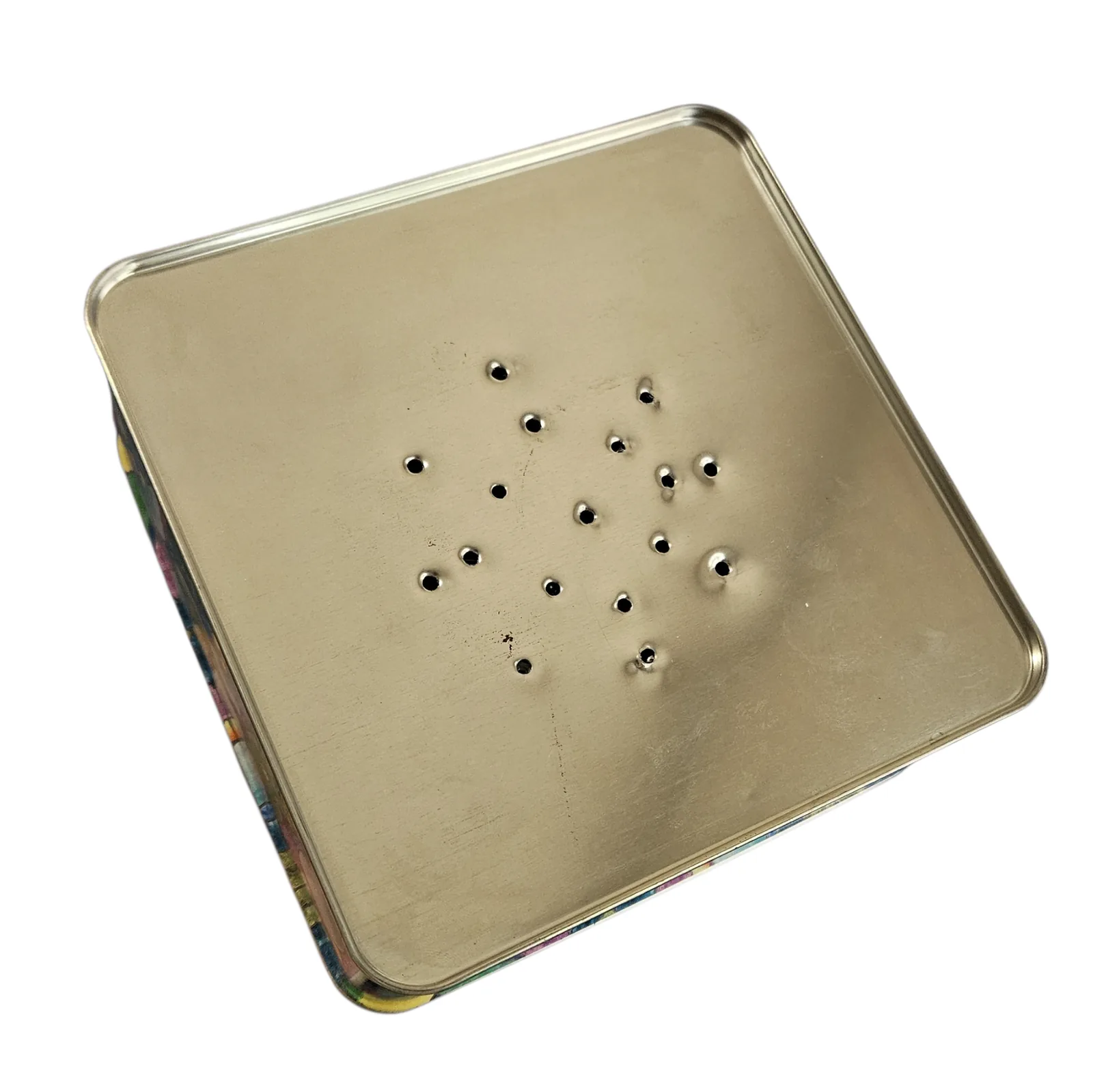 The plain metal underside of the tin, seen at an angle against white, with a loose cluster of about twenty small holes drilled through the center of it. The printed pansy pattern of the sides shows along the near edges.