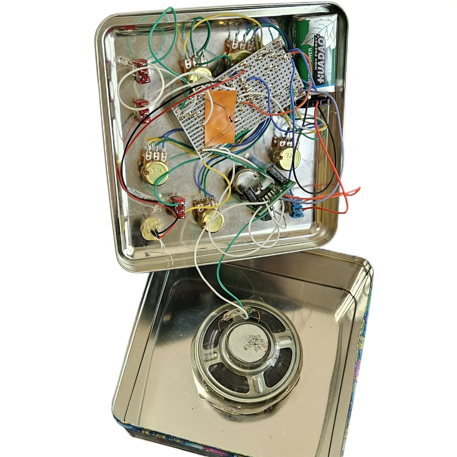 The tin open against white, its lid hinged up to show the underside of the panel and the base below. On the lid: a rectangular stripboard mounted in the middle with an orange square of tape over one patch, six gold potentiometer bodies and several small red toggle-switch bodies fixed through the metal, a small green amplifier module wired in near the lower right, a green 9V battery in the top right corner, and a tangle of red, black, white, green, blue, yellow and orange leads between them. In the base: a round speaker glued face down in the center of the plain metal, its magnet upward, joined to the lid by a white and a green wire.