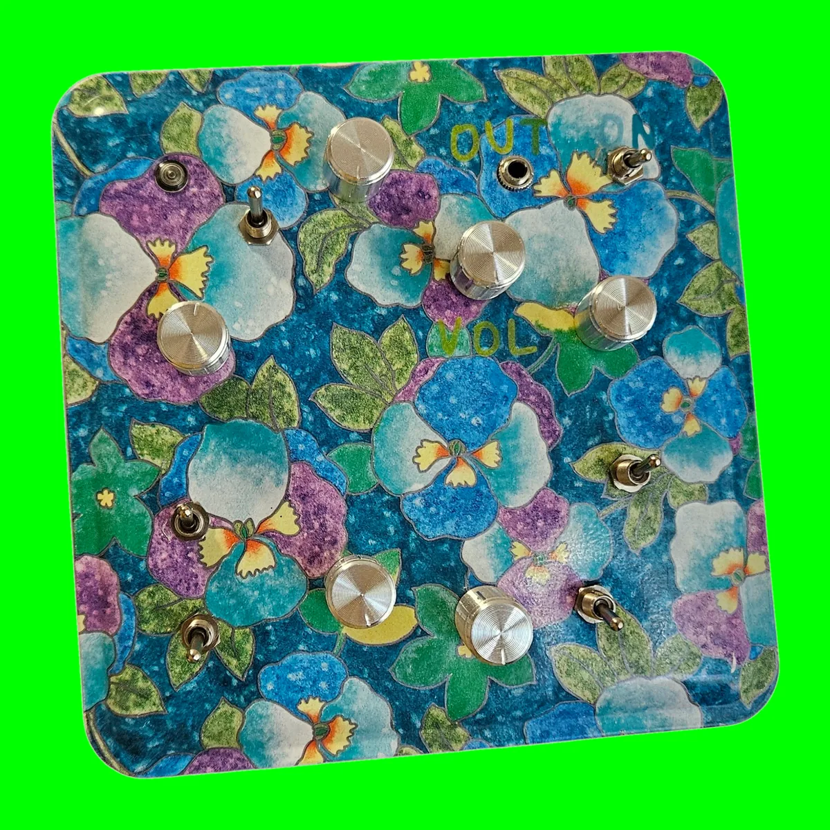 Garden Variety - a square tin lid printed all over with blue, purple and white pansies, with six brass knobs, six brass toggle switches, a jack socket and a small indicator lamp set into it, and OUT, ON and VOL hand-lettered beside them