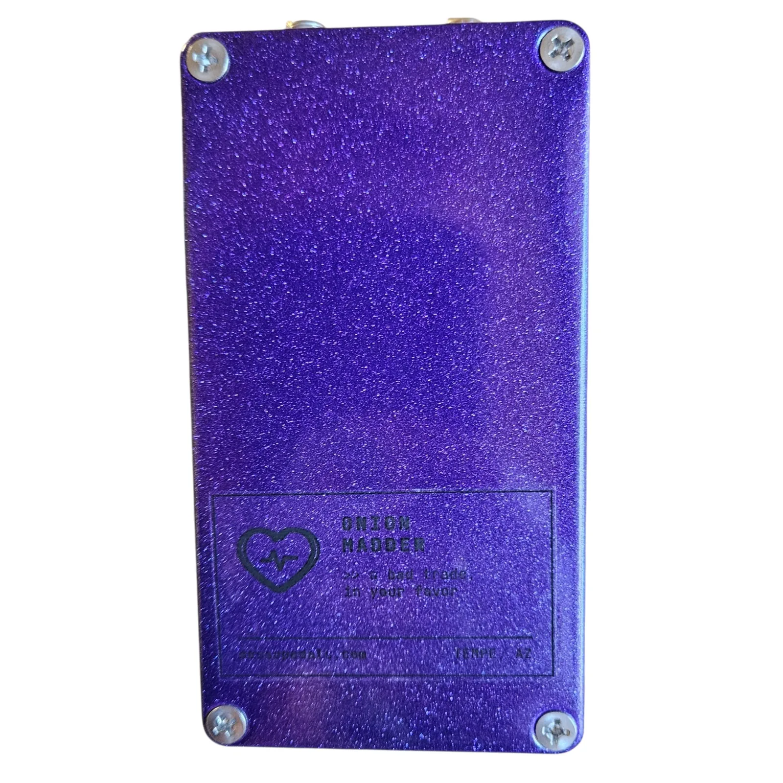 Get Wet pedal, back view showing the metallic-purple-glitter bottom plate with a Phillips screw in each corner and a label printed in black near the bottom: a heart with a heartbeat line, ONION MADDER, a bad trade, in your favor, messopedals.com and TEMPE, AZ.