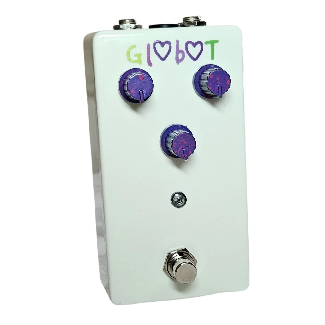 A white pedal photographed face on. The name is hand-painted across the top as Gl<3b<3T, with a purple heart standing in for each of the two O's, the letters in green and purple. Three purple glitter knobs sit below it, two side by side and one centered beneath them, with a small LED and a silver footswitch under those.