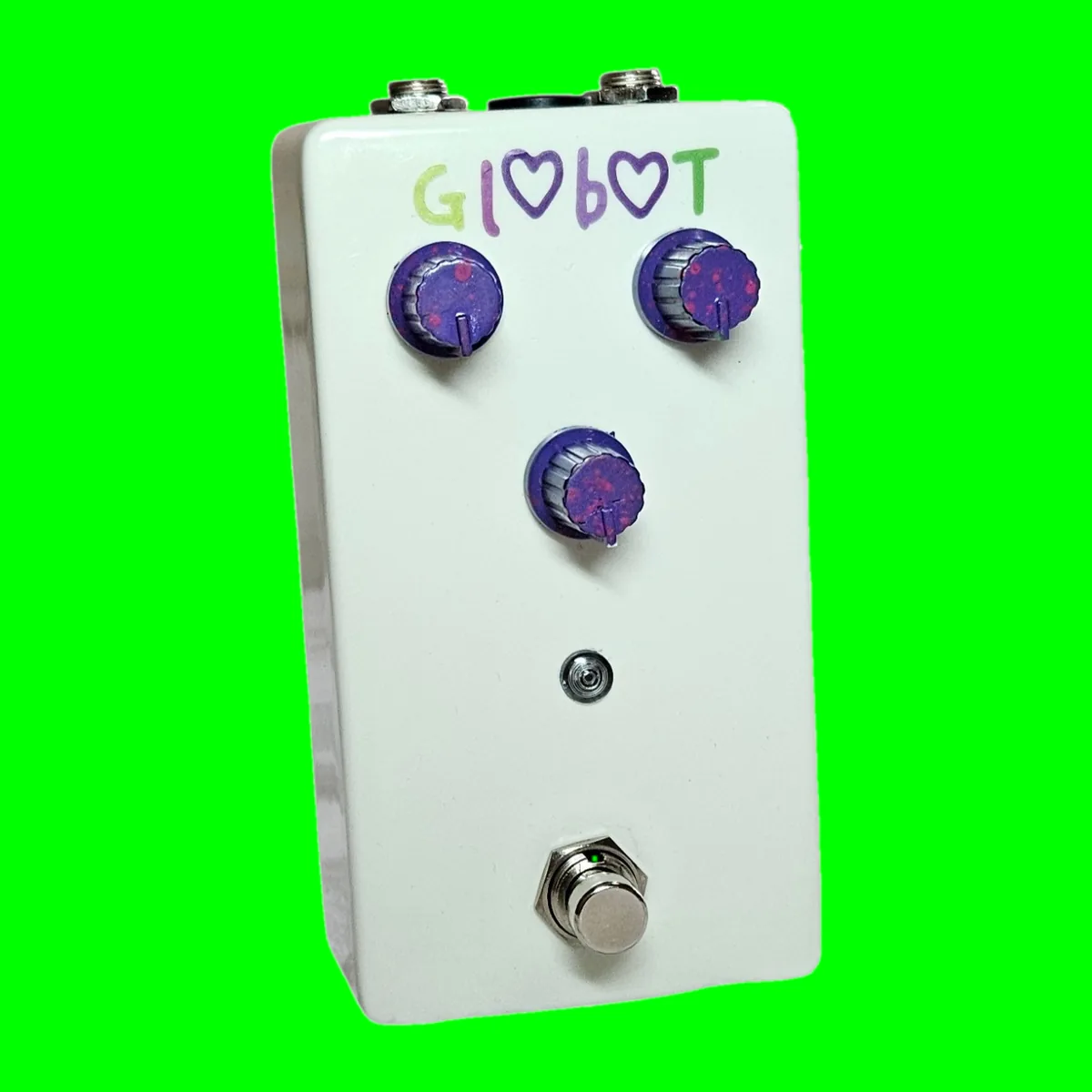 A white pedal photographed face on. The name is hand-painted across the top as Gl<3b<3T, with a purple heart standing in for each of the two O's, the letters in green and purple. Three purple glitter knobs sit below it, two side by side and one centered beneath them, with a small LED and a silver footswitch under those.