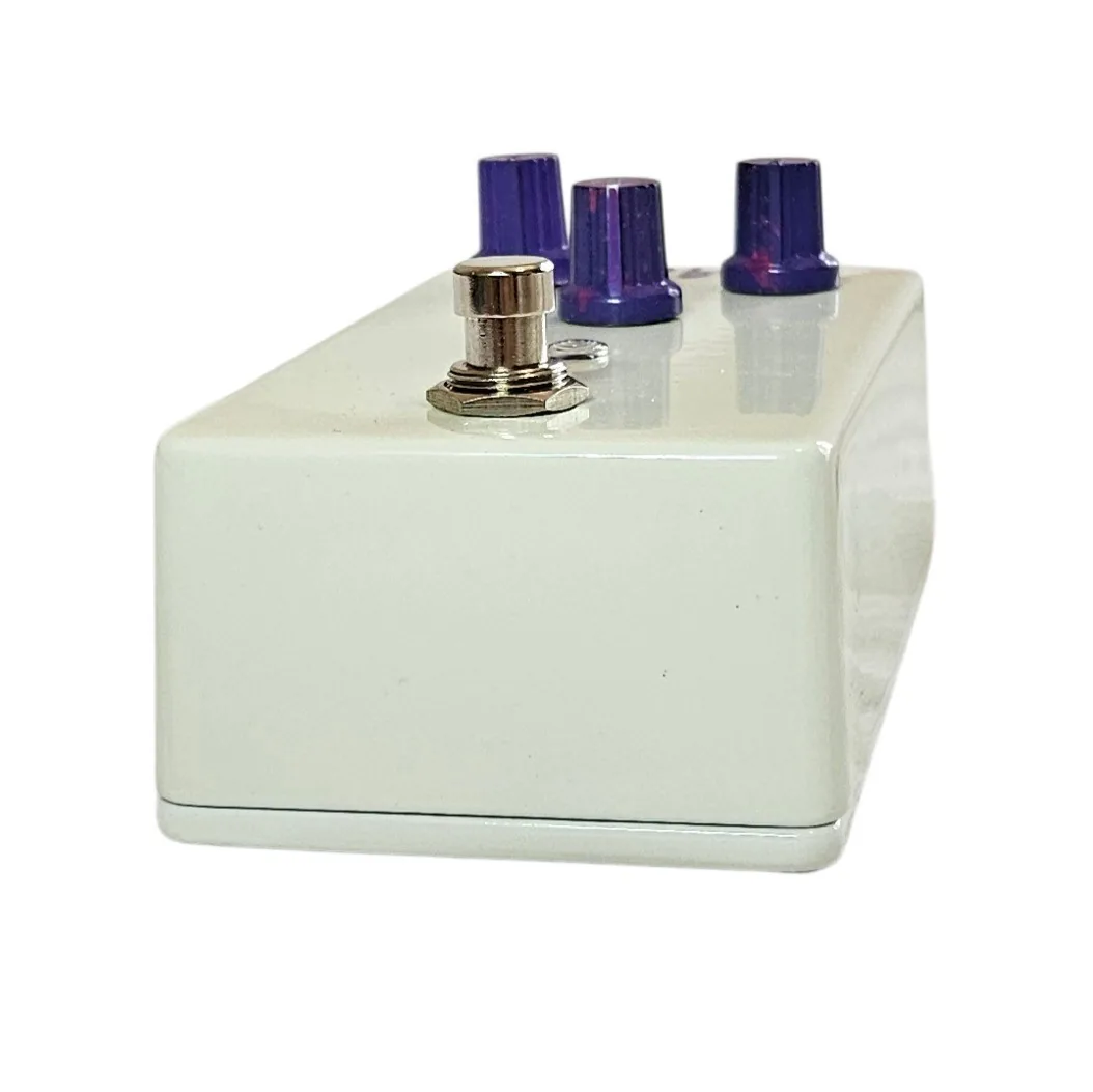 Looking down on the pedal from the footswitch end: the three purple glitter knobs stand along the far edge, the silver footswitch nut is in the foreground, and the plain white wall of the enclosure fills the lower half.