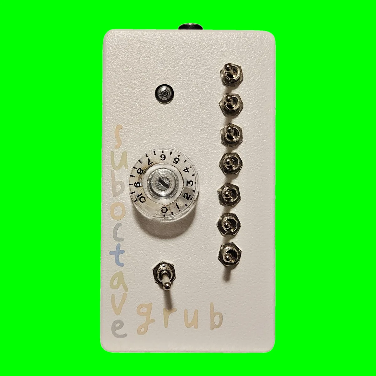 Sub Octave Grub - an upright off-white box with seven chrome toggle switches down the right side, a clear numbered knob and an LED on the face, an eighth toggle below and the words sub octave down the left edge with grub across the bottom in pastel hand lettering, on a chroma green tile