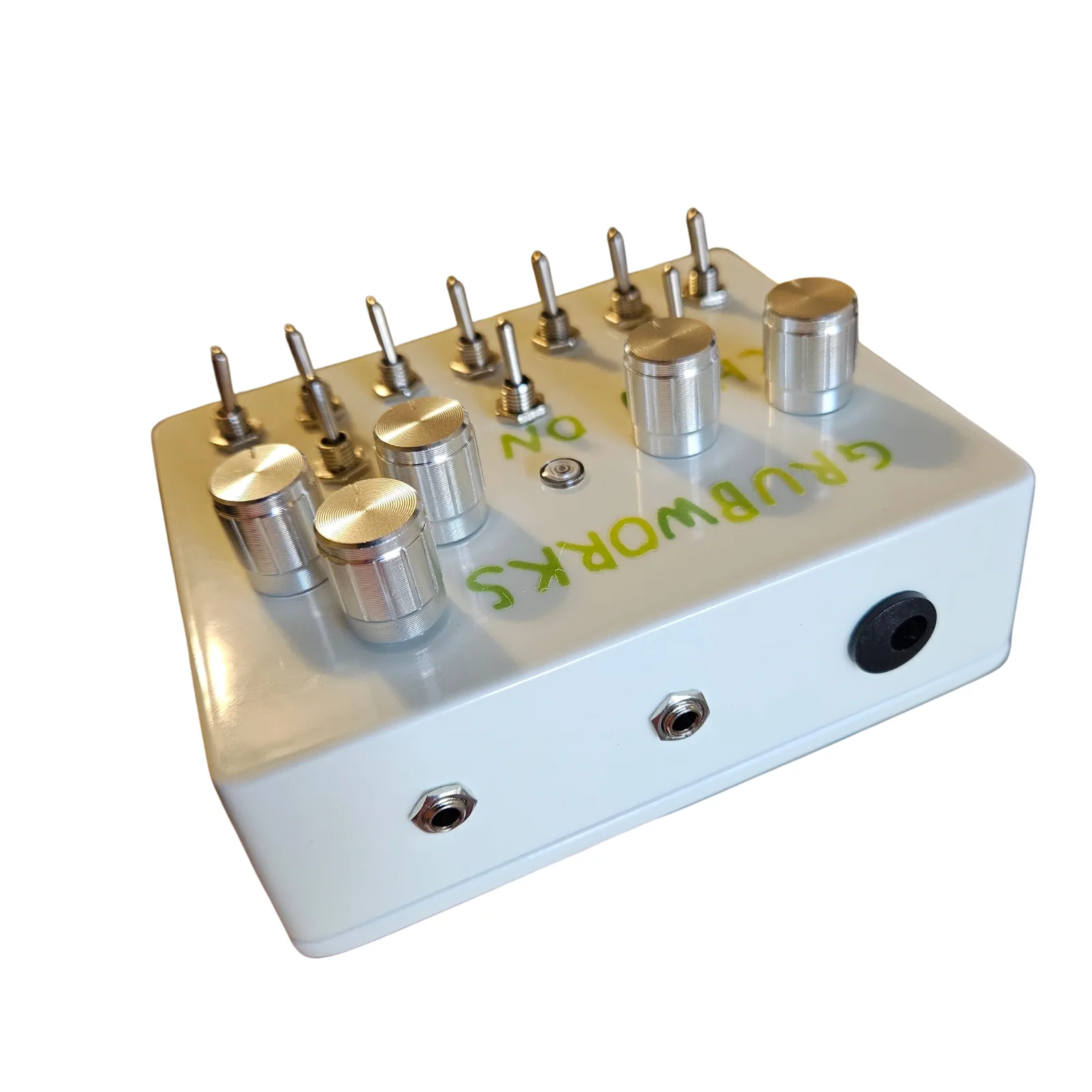 The box seen from its top edge at a low angle, GRUBWORKS upside down on the face beyond: two small jacks along the edge and a round black power socket at the right end, with the knobs and toggles standing up behind.
