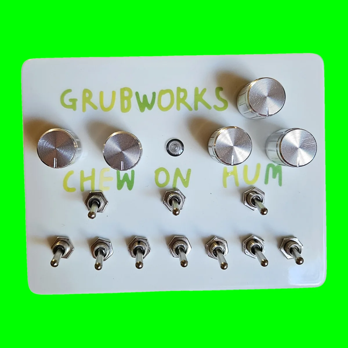 A glossy white box photographed face on, GRUBWORKS hand-lettered across the top in green, with a chrome knob on its own at the top right. Below the name is a row of four chrome knobs, two at the left and two at the right, with a small LED between them. CHEW, ON and HUM are lettered across the middle, each over a toggle, and a row of seven toggles runs along the bottom edge.