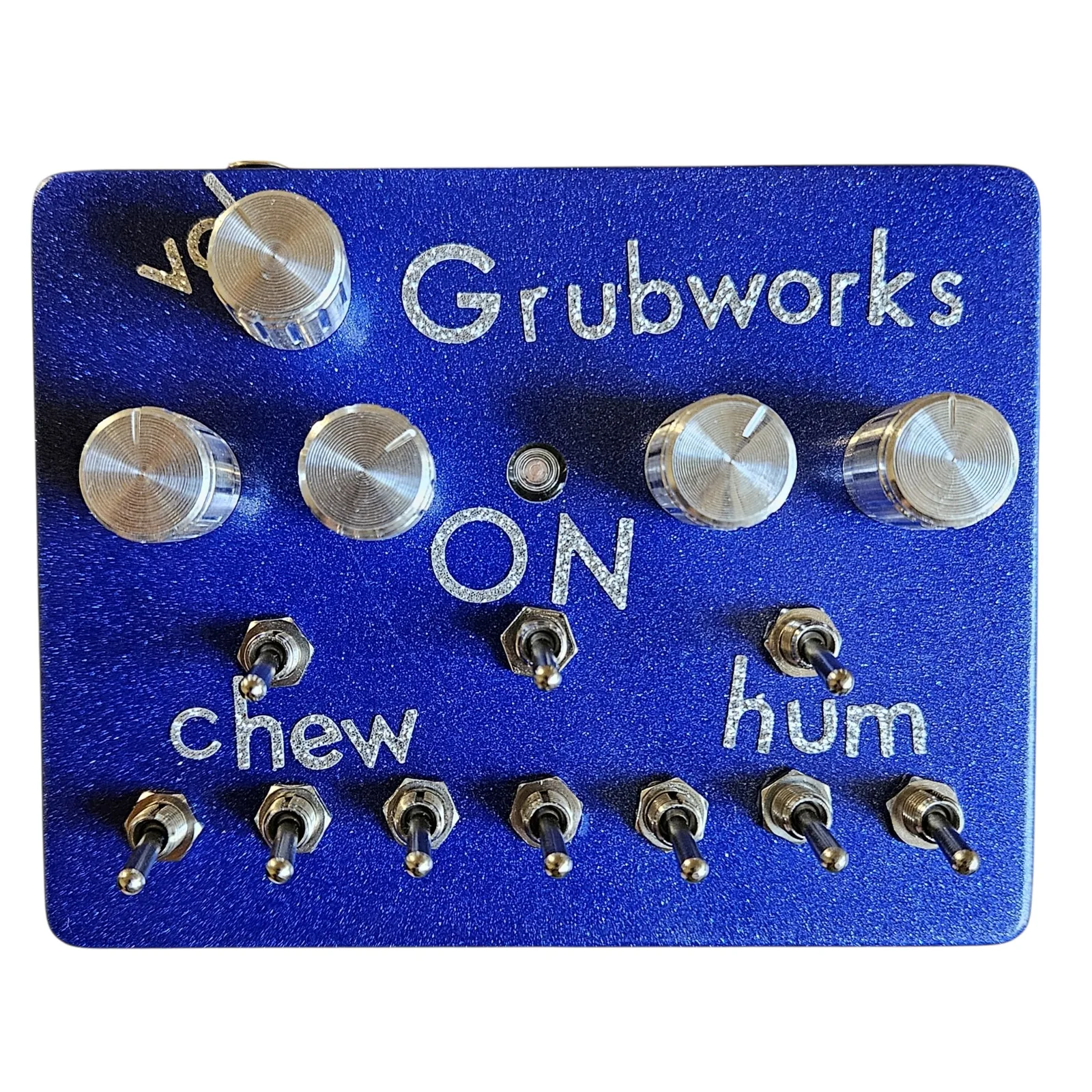 A metallic blue glitter box photographed face on. Grubworks is lettered across the top in sparkly silver, with vol beside a chrome knob at the top left. Below that is a row of four chrome knobs, two at the left and two at the right, with a small LED between them and ON lettered under the LED. Three toggles sit under the knobs, one over the word chew at the left, one under ON in the middle and one over the word hum at the right, and a row of seven toggles runs along the bottom edge.