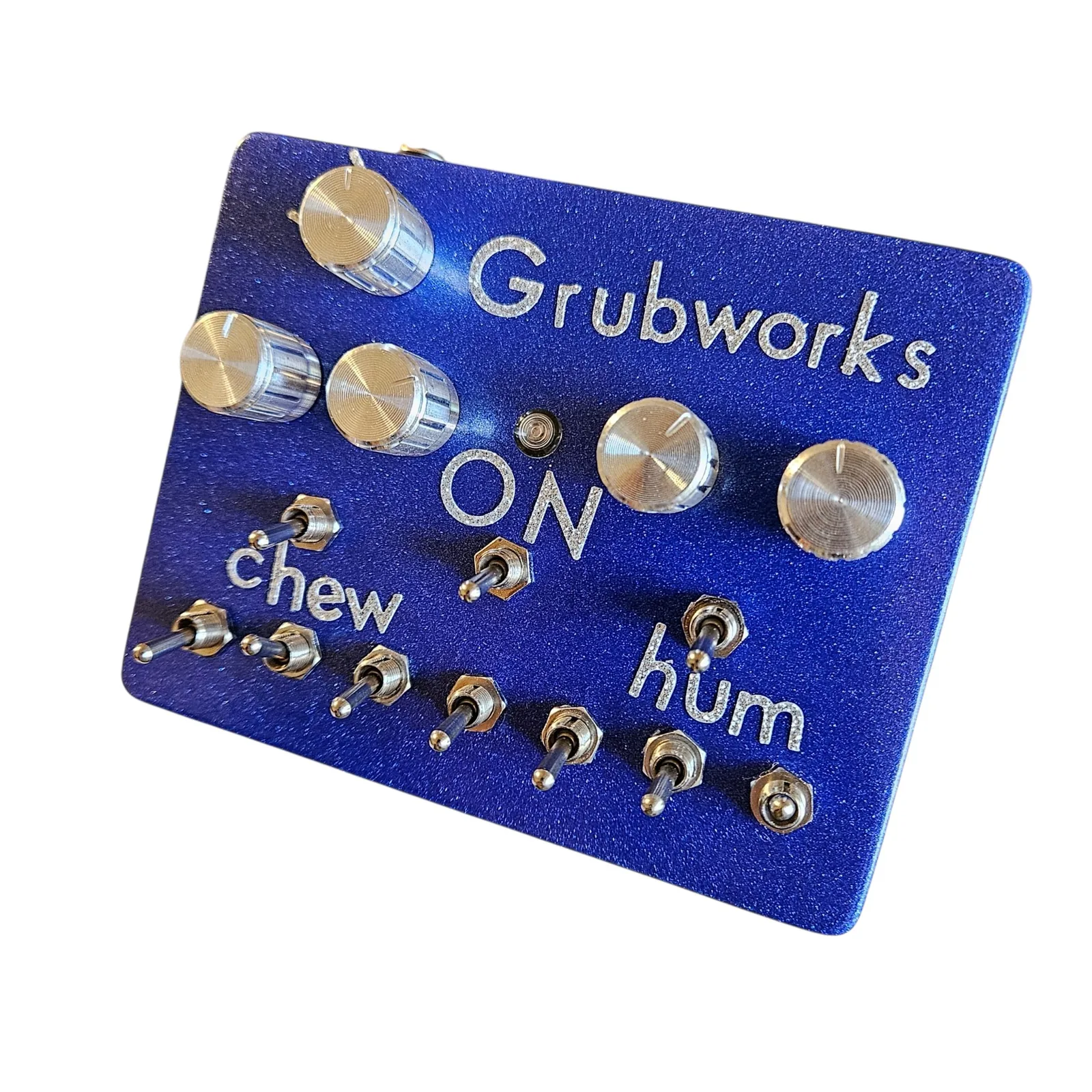 The box tilted from its left to show the face: vol and its knob at the top left, Grubworks, the four chrome knobs with the LED and ON between them, chew and hum over their toggles, and the row of seven toggles along the bottom edge.