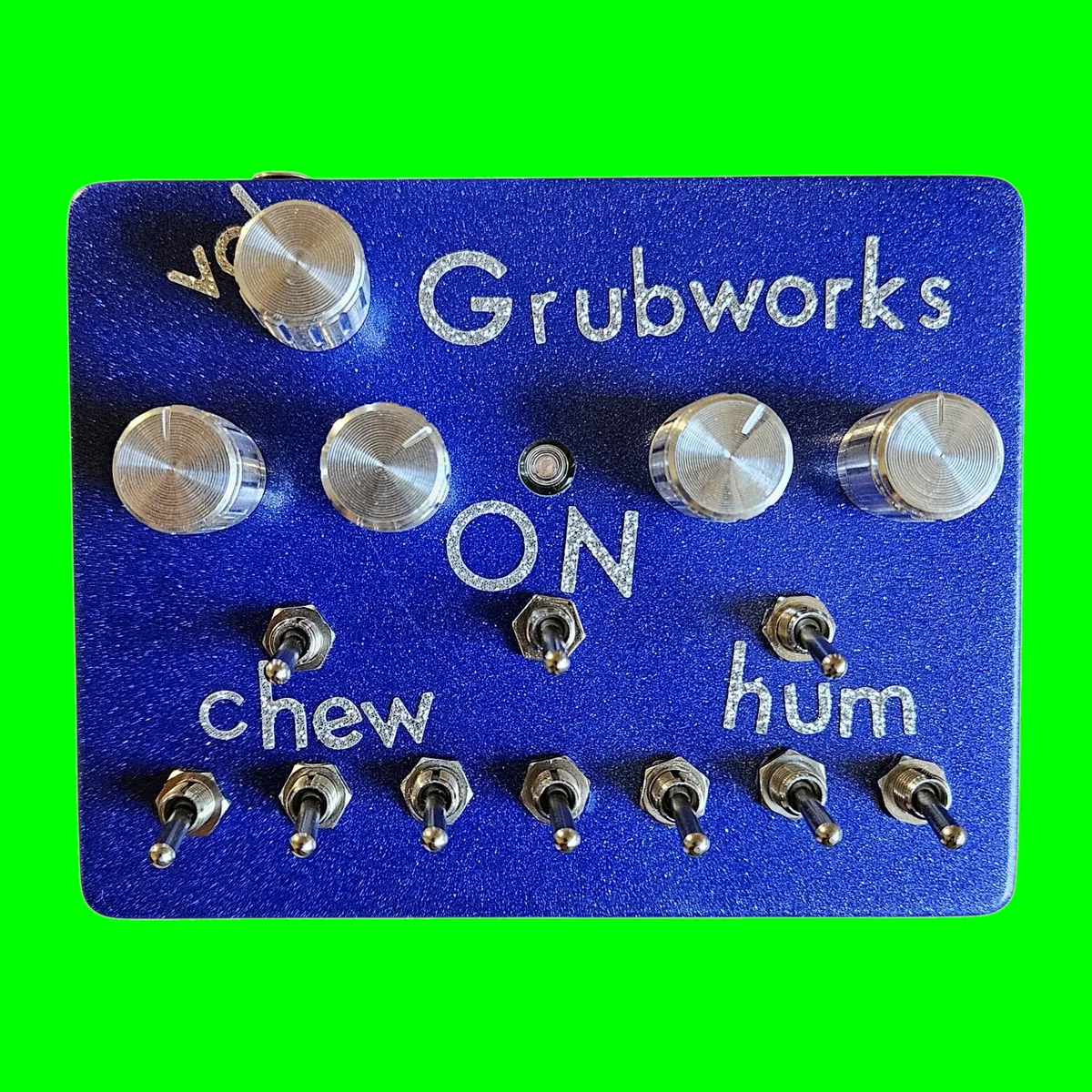 A metallic blue glitter box photographed face on. Grubworks is lettered across the top in sparkly silver, with vol beside a chrome knob at the top left. Below that is a row of four chrome knobs, two at the left and two at the right, with a small LED between them and ON lettered under the LED. Three toggles sit under the knobs, one over the word chew at the left, one under ON in the middle and one over the word hum at the right, and a row of seven toggles runs along the bottom edge.