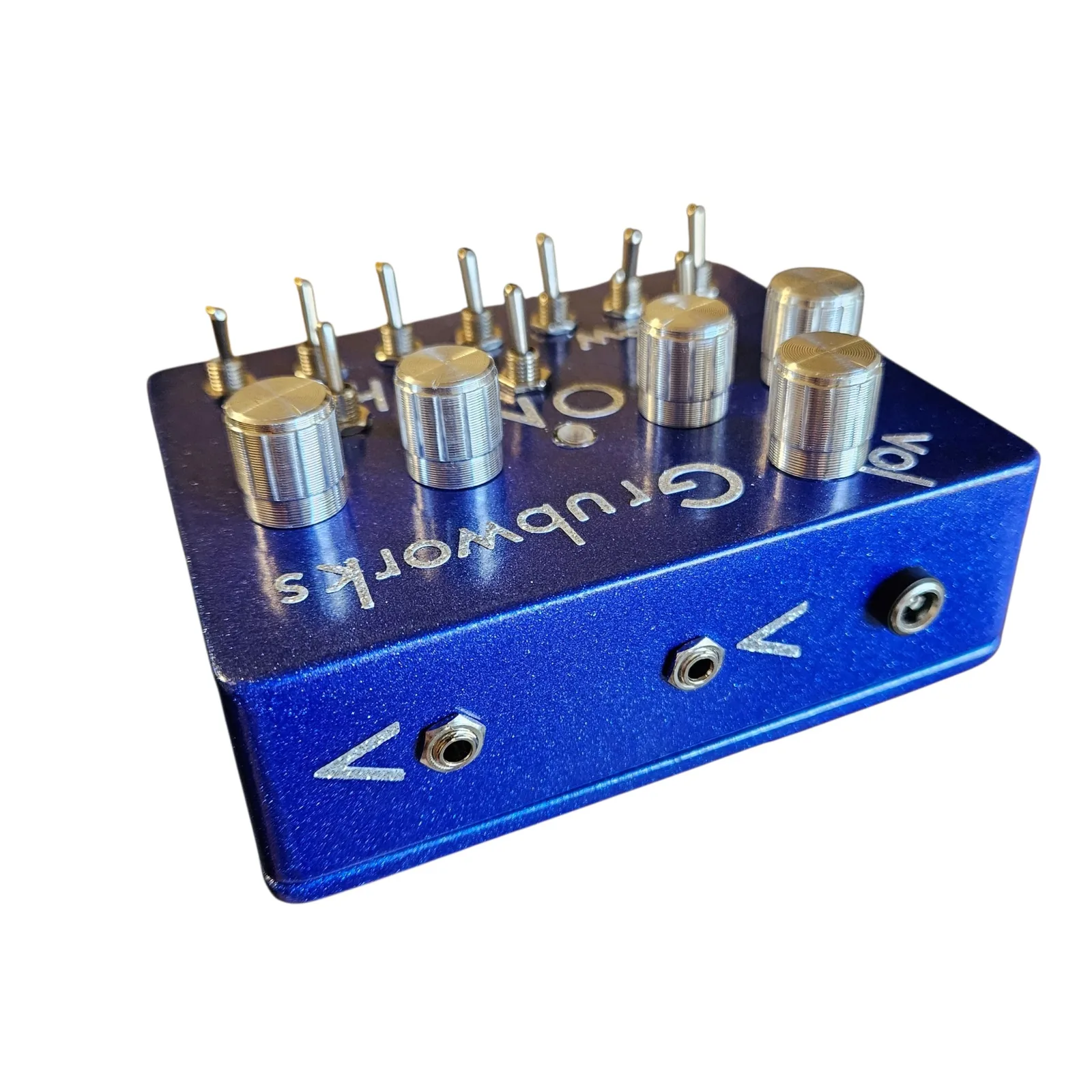 The top edge of the box facing the camera, with the knobs and toggles standing up beyond it and Grubworks and vol lettered upside down on the face. Two jacks sit along the edge with a silver arrow drawn beside each, and a third socket is at the right end.
