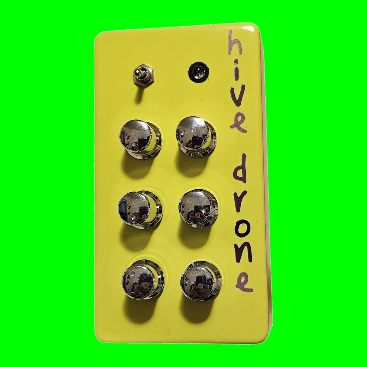 Hive Drone - an upright neon green box with six chrome knobs in two columns of three, a toggle switch and an LED at the top and the name hand-lettered in purple down the side, on a chroma green tile