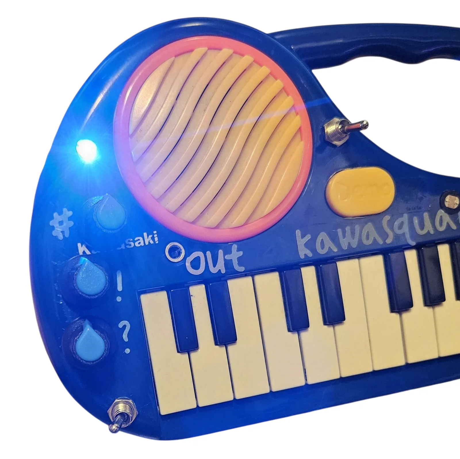 The same corner with the oscillator running instead. The indicator is blazing a bright blue-white, bright enough to flare across the lens and wash the whole blue panel and the orange speaker ring in colored light. The toggle beside the speaker is flipped down.