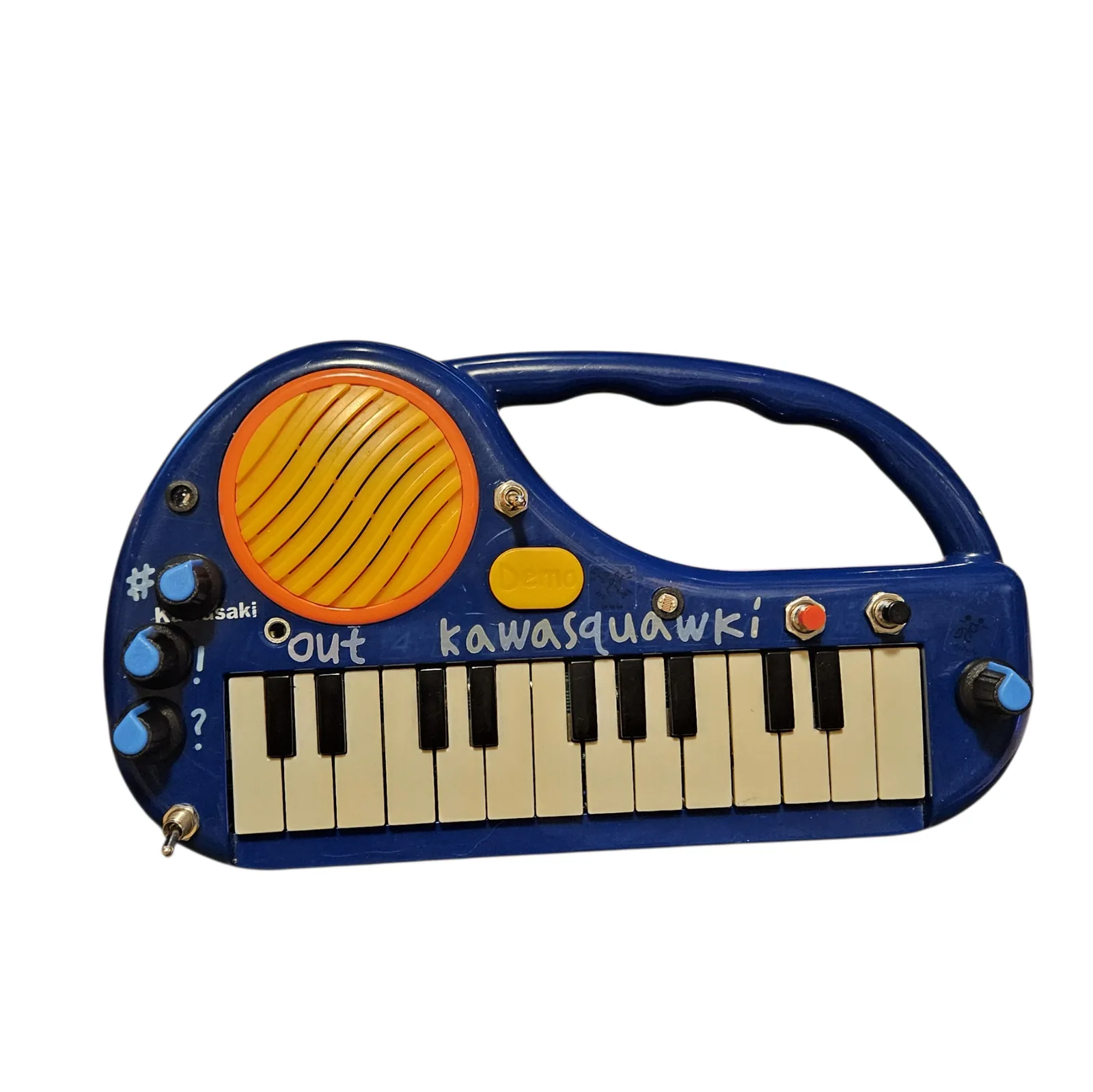 The Kawasquawki head on against white: a dark blue toy keyboard with a rounded teardrop body and a molded carry handle at the top right. A big round speaker grille of yellow bars inside an orange ring fills the top left, with a small yellow Demo button beside it, and twenty-five small keys run along the bottom. kawasquawki is hand-lettered across the panel in pale blue, and out is lettered beside a small jack. Three black knobs with blue teardrop pointers run down the left edge, marked with an asterisk, an exclamation mark and a question mark; a fourth is at the far right. Between them sit a red button, a black button, a photocell, and toggle switches at the top left, beside the speaker and at the bottom left corner.