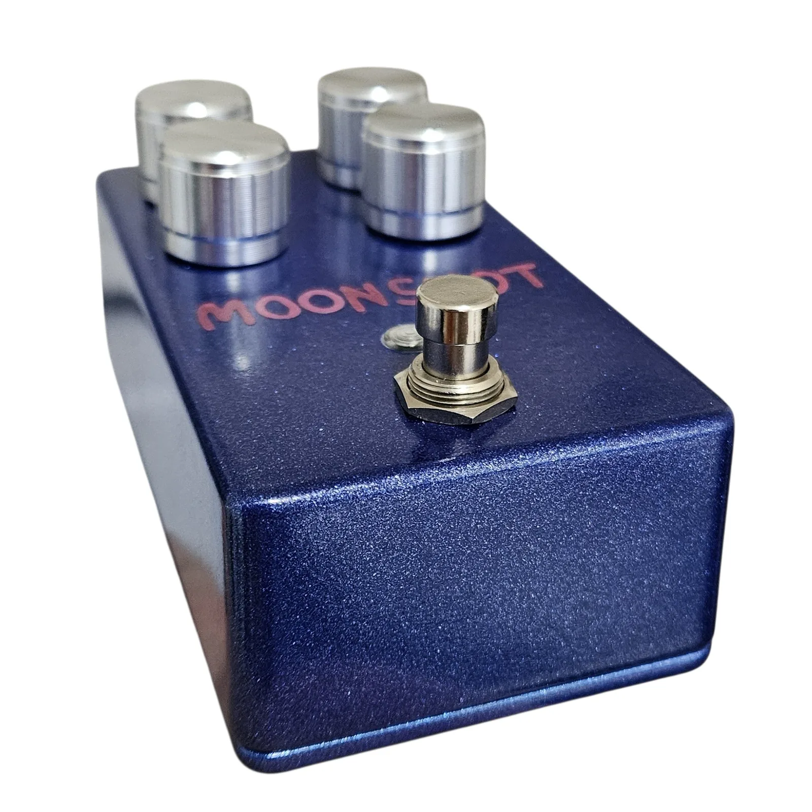 A low angle from the footswitch end, looking along the blue sparkle enclosure past the silver footswitch and the red lettering to the four knobs at the far end.