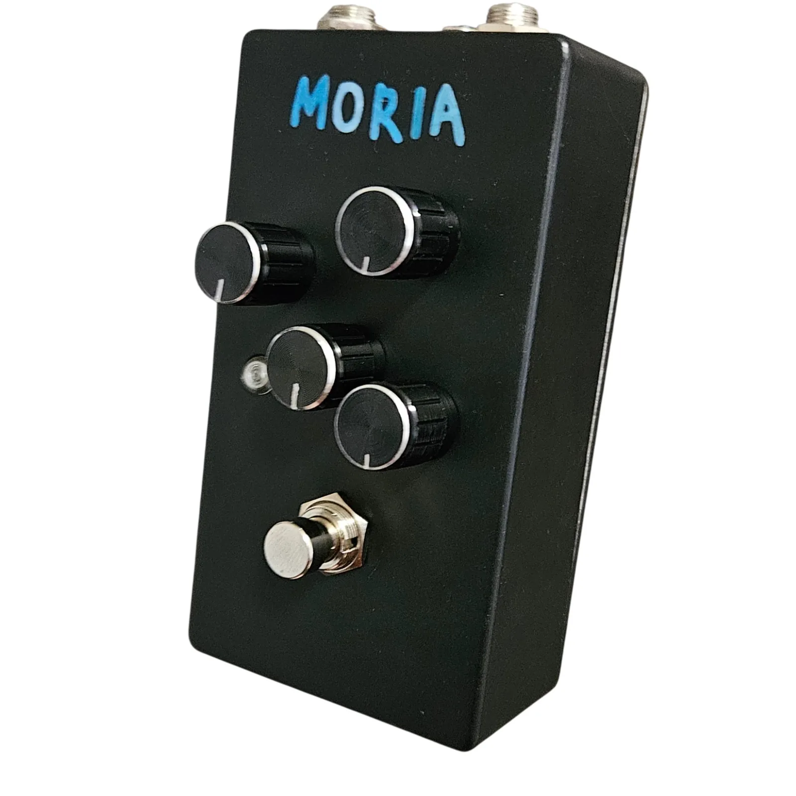 A black upright pedal at a three-quarter angle, with MORIA written across the top in blue marker in blocky capitals. Four black knurled knobs with silver caps and white pointer lines stand on the face in a staggered pattern, two upper and two lower. A small round LED bezel sits at the left between the lower pair, and a silver footswitch is at the bottom. Two quarter-inch jack nuts and a smaller barrel jack show along the top edge.