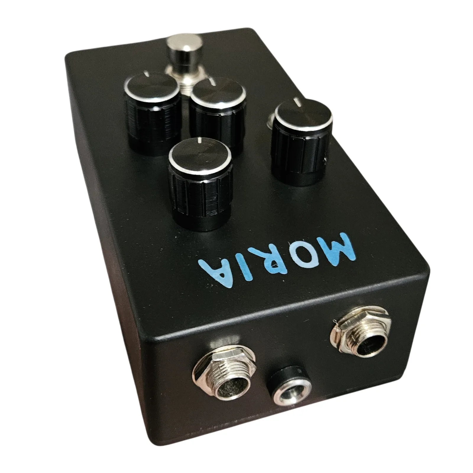 The pedal tipped forward so its top edge faces the camera. A quarter-inch jack sits at each end of the edge with a black barrel jack for the power supply between them, set lower. The face falls away below, with MORIA reading mirrored from this angle and the four knobs beyond it.