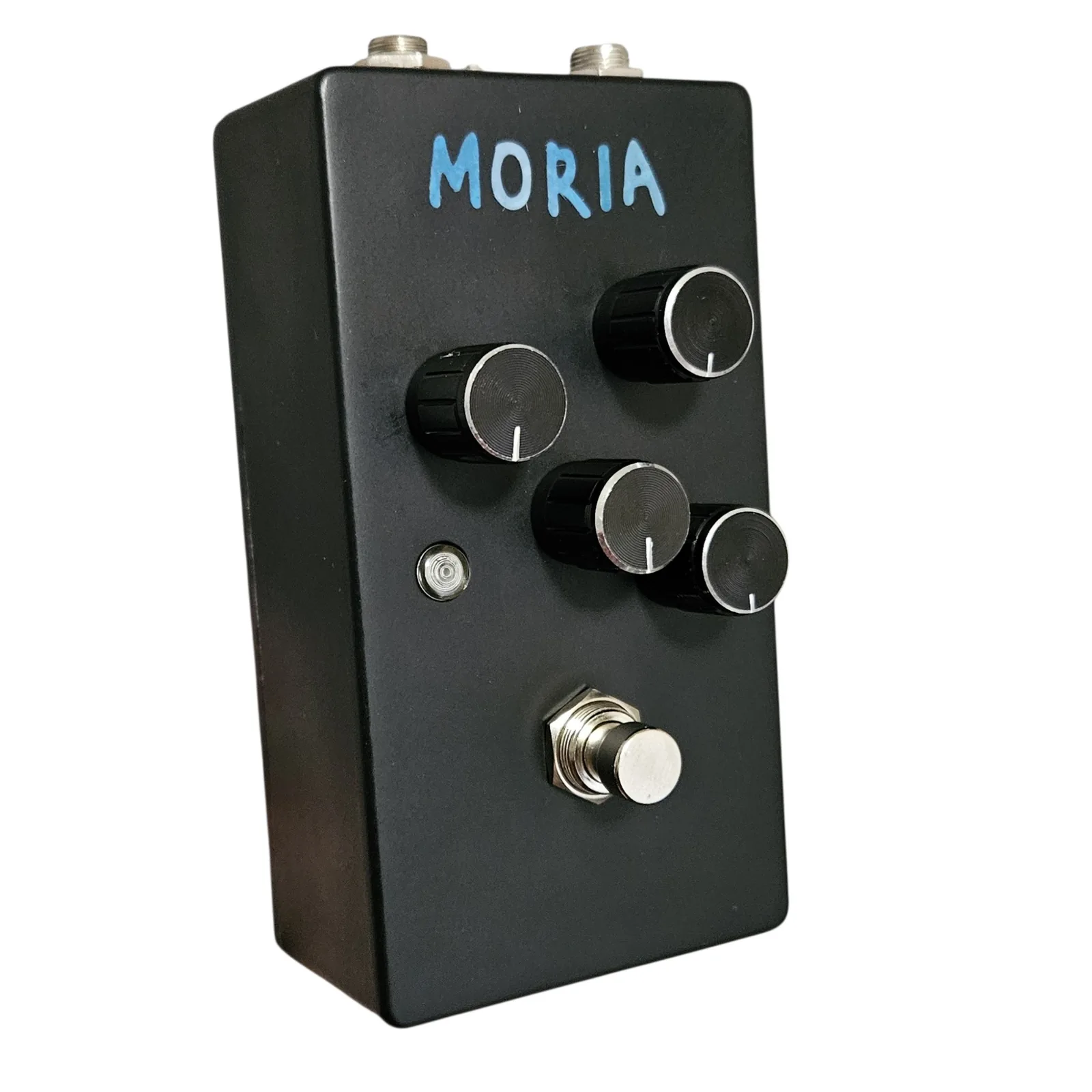 The same black pedal turned three quarters the other way, so the left flank and the face are both in view. The four knobs and the footswitch stand off the face in profile and MORIA reads across the top in blue. The flank is plain black with nothing on it.