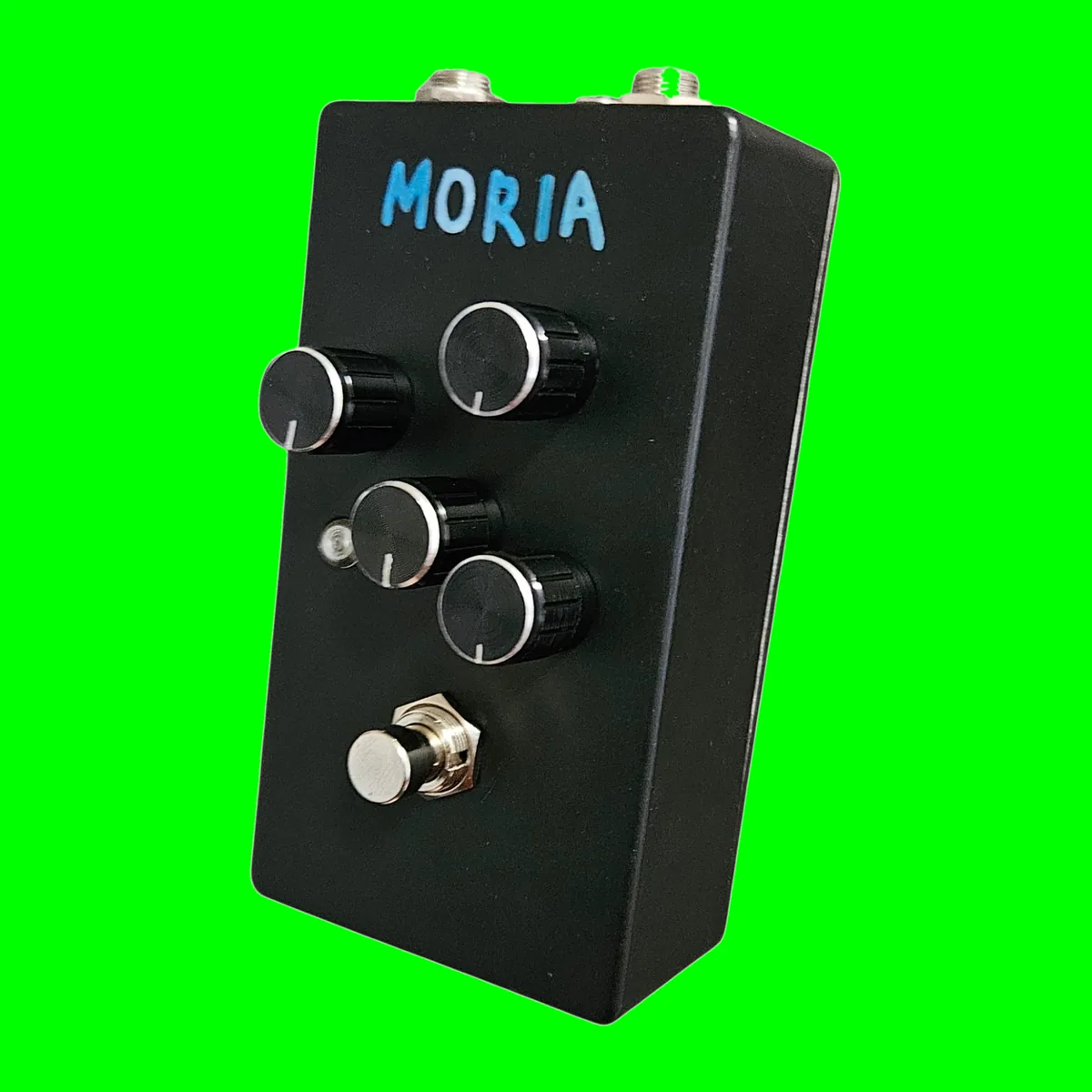 A black upright pedal at a three-quarter angle, with MORIA written across the top in blue marker in blocky capitals. Four black knurled knobs with silver caps and white pointer lines stand on the face in a staggered pattern, two upper and two lower. A small round LED bezel sits at the left between the lower pair, and a silver footswitch is at the bottom. Two quarter-inch jack nuts and a smaller barrel jack show along the top edge.