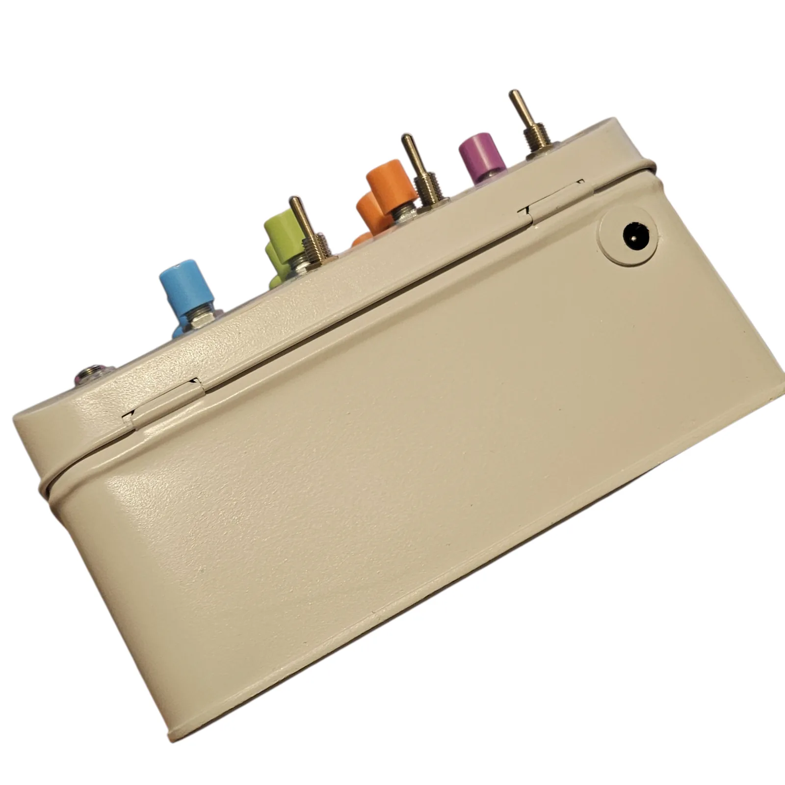 The tin tipped over so one long painted side faces the camera. A single black barrel socket is set into the side wall near one end, and the knobs and toggle levers stick out along the far edge of the lid.