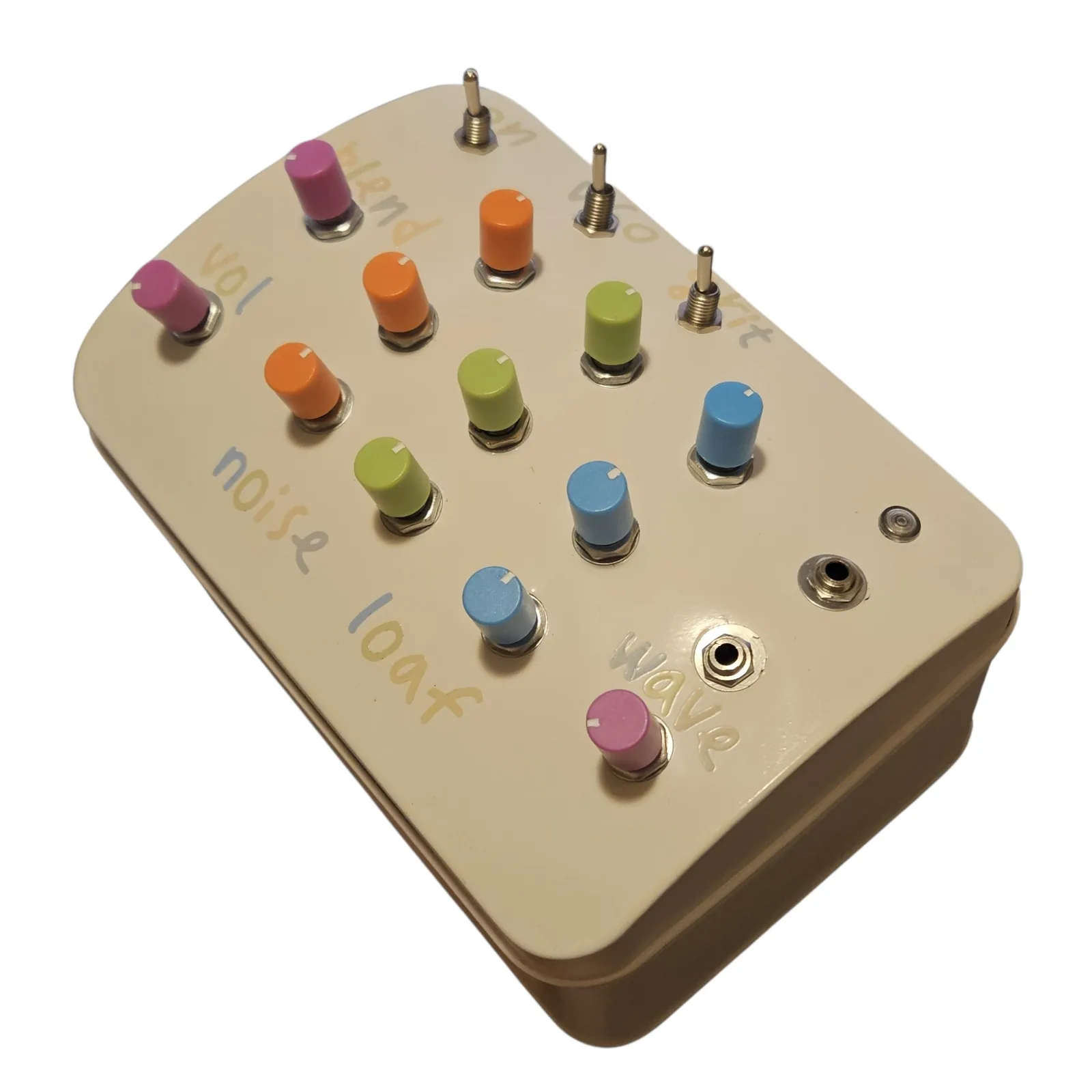 The tin at a three-quarter angle with the lettered corner nearest the camera. Two small silver sockets and an unlit LED sit in the cream lid near the corner marked wave, with the twelve colored knobs and the three toggles standing up behind them and the plain painted side of the tin below.
