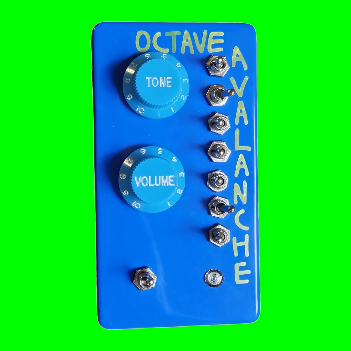 An upright glossy blue box photographed face on, OCTAVE hand-lettered in pale green across the top and AVALANCHE running down the right edge. Two light blue knobs with numbered skirts, lettered TONE and VOLUME, sit one above the other on the left, seven chrome toggle switches run down the right, and at the bottom there is a toggle on the left and a small round bezel on the right.