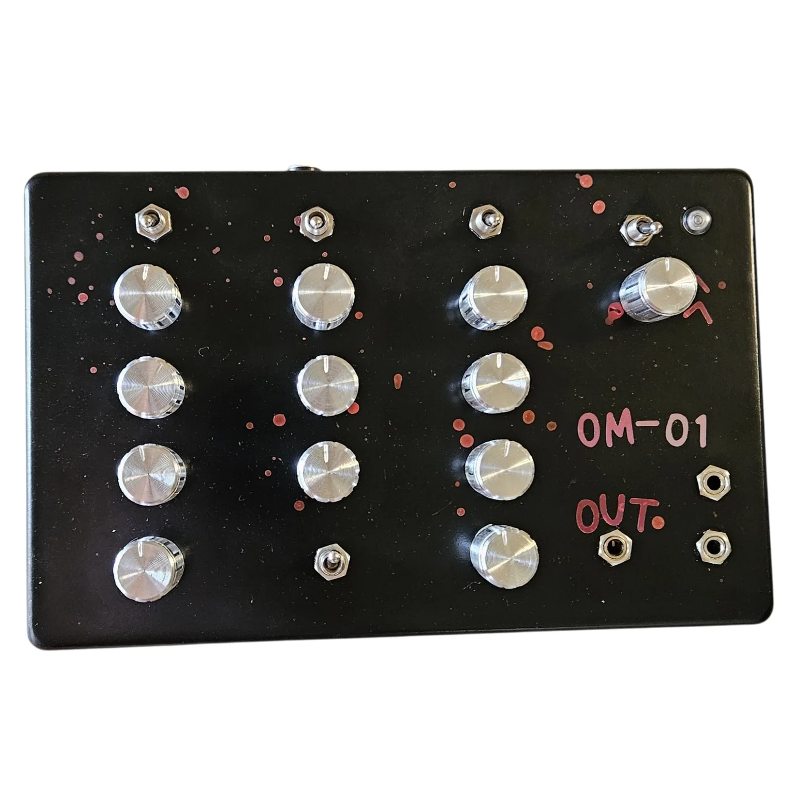 A wide black enclosure photographed face on, with OM-01 hand-lettered in pink marker at the right and OUT written below it. Twelve silver knurled aluminum knobs stand in four columns: four in the first, three in the second, four in the third, and one larger knob on its own at the top right. Four small silver toggles run across the top of the face and a fifth sits low in the second column. A round indicator lamp is set in the top right corner. Three small jack sockets are grouped at the bottom right, one of them directly under the word OUT, and the nut of a barrel power jack shows on the top edge.