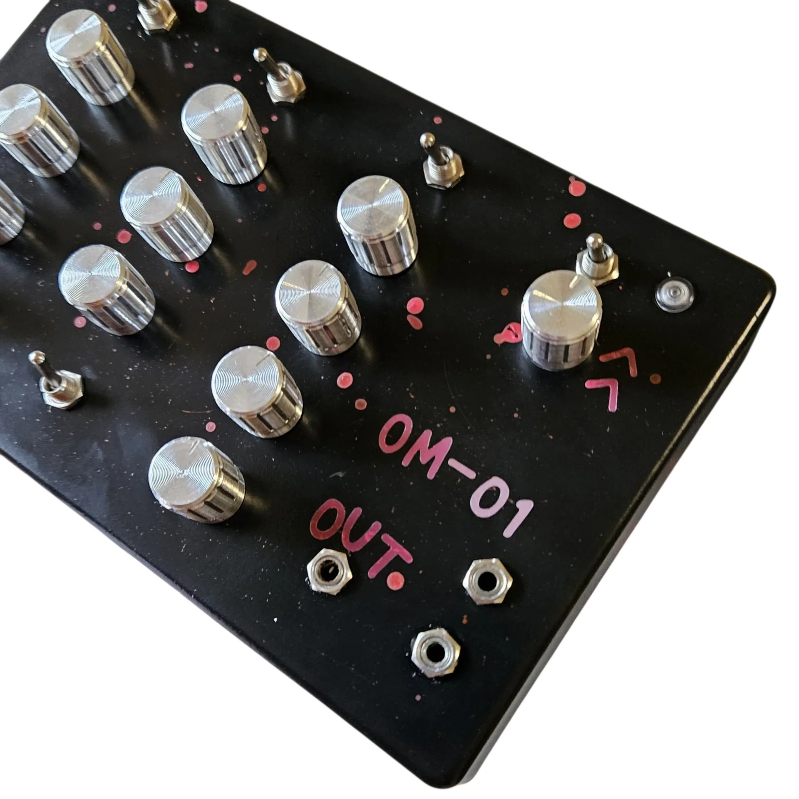 A close three-quarter of the right-hand end of the face. The larger silver knob sits below its own toggle with a round indicator lamp beside it in the corner, and OM-01 and OUT are hand-lettered in pink below. Three small silver jack sockets are grouped in the bottom corner. The knobs are knurled aluminum with a white pointer line down the side.