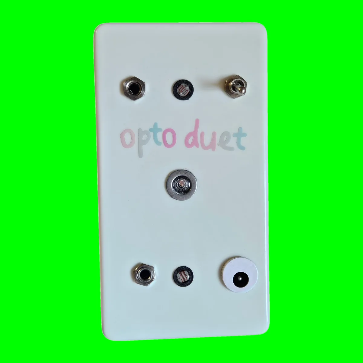 Opto Duet - an upright white glow-in-the-dark box with opto duet lettered in pastel across the top, a jack, a photocell and a toggle in a row above the name, a round LED bezel in the middle, and a second jack, a second photocell and a DC socket along the bottom