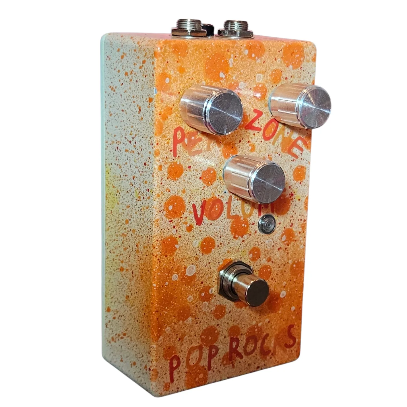 Left side of the pedal: a plain cream wall flecked with orange and red, showing the three pink barrel knobs standing off the face, a small LED beside the Volume knob, and the footswitch mounted on a white washer.
