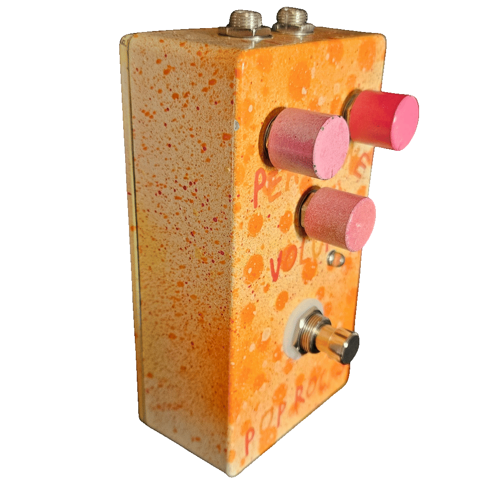Left side of the pedal: a plain cream wall flecked with orange and red, showing the three pink barrel knobs standing off the face, a small LED beside the Volume knob, and the footswitch mounted on a white washer.