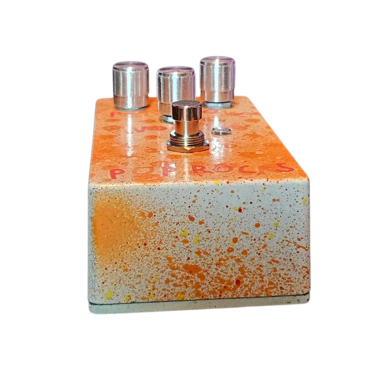 Top-down view of the enclosure: hand-lettered orange "IN" marks the left 1/4-inch jack and "OUT" the right, with a DC power jack set between and slightly behind them. The three pink knobs and the footswitch fall away below.