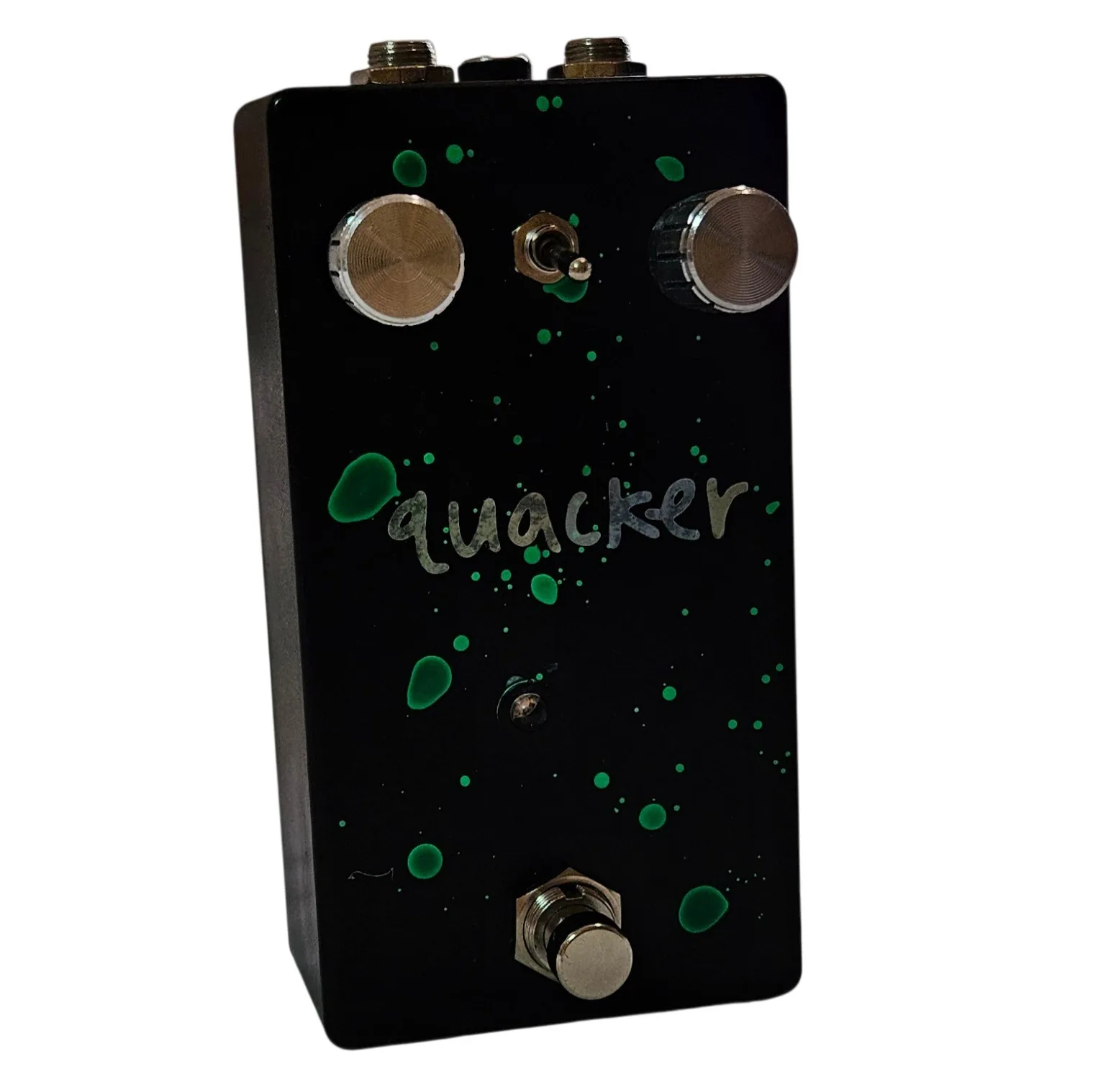 A tall black pedal photographed face on, flecked all over with hand-flicked green paint spatters. The word quacker is painted across the middle in silver lowercase script. Two knurled silver knobs sit at the top corners with a small toggle switch between them, a tiny LED sits below the lettering, and a silver footswitch stands at the bottom.