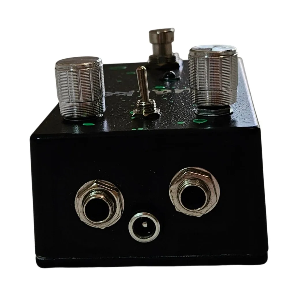 The pedal's top edge straight on, with two silver quarter-inch jack sockets and a barrel power socket set low between them. The knobs and toggle switch are visible above, and green flecks dot the black.