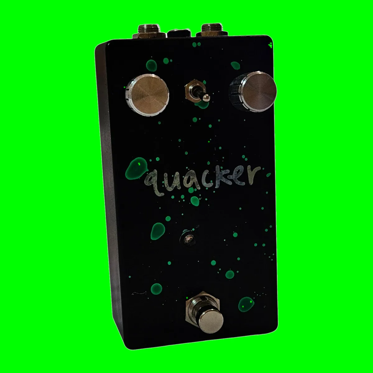 A tall black pedal photographed face on, flecked all over with hand-flicked green paint spatters. The word quacker is painted across the middle in silver lowercase script. Two knurled silver knobs sit at the top corners with a small toggle switch between them, a tiny LED sits below the lettering, and a silver footswitch stands at the bottom.