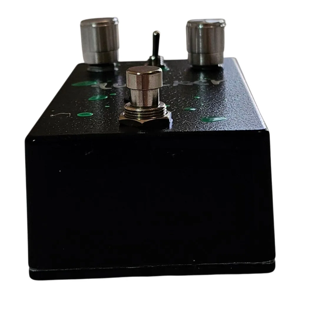 Looking down on the pedal's top edge, showing two knurled silver knobs either side of a small metal toggle switch, with green paint flecks across the black between them and the footswitch below.